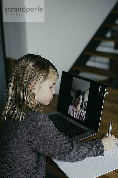 Mädchen schreibt auf Papier und lernt zu Hause per Videoanruf am Laptop