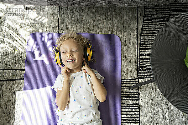 Fröhliches Mädchen  das Musik über Kopfhörer genießt und zu Hause auf einer Trainingsmatte liegt