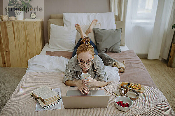Frau beim E-Learning per Laptop  liegt mit Kaffeetasse neben Hund zu Hause im Bett