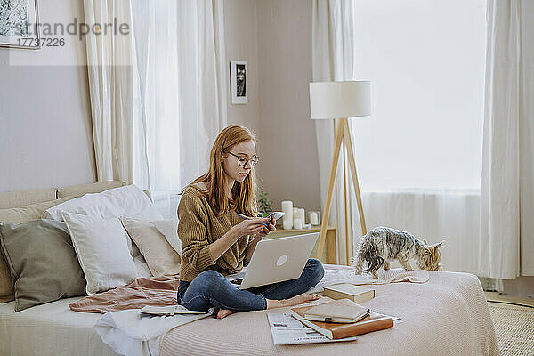 Frau sitzt mit Laptop und Hund im heimischen Bett und benutzt Smartphone