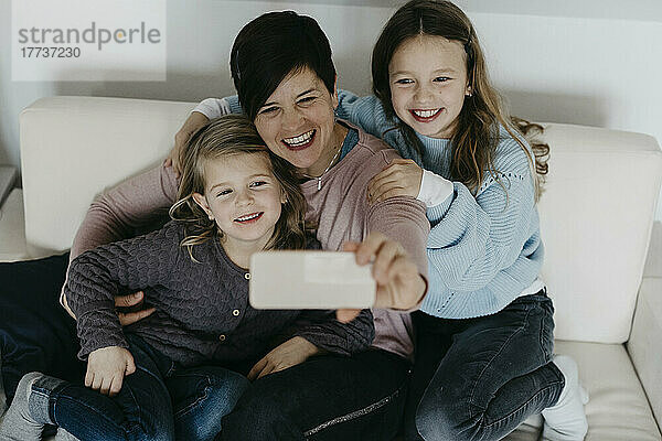 Lächelnde Mutter macht Selfie mit Töchtern  die zu Hause auf dem Sofa sitzen