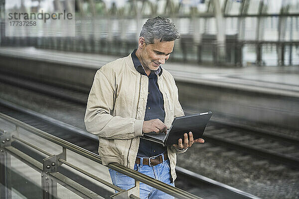 Lächelnder Geschäftsmann mit Tablet-PC  der am Bahnhof steht