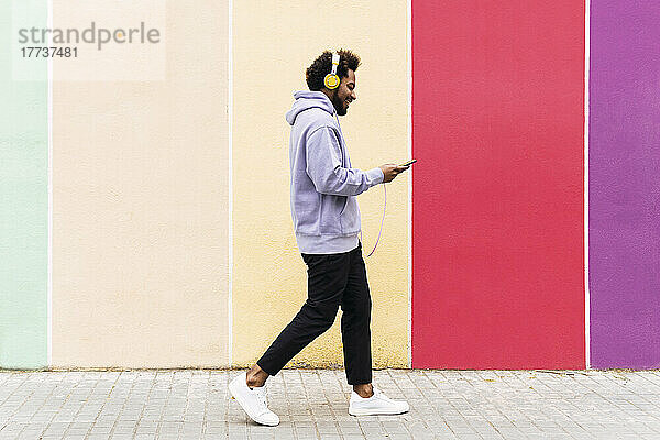 Lächelnder Mann  der sein Mobiltelefon benutzt und über Kopfhörer Musik hört  während er auf dem Fußweg läuft
