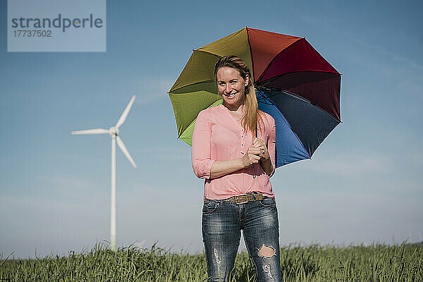 Lächelnde Frau steht an einem sonnigen Tag mit buntem Regenschirm im Feld