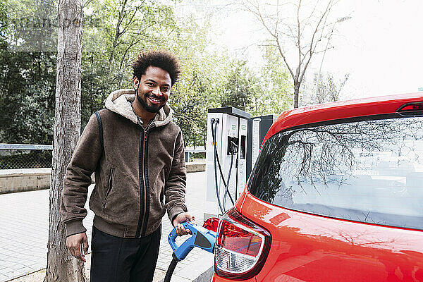 Lächelnder Afro-Mann lädt am Bahnhof ein rotes Elektroauto auf