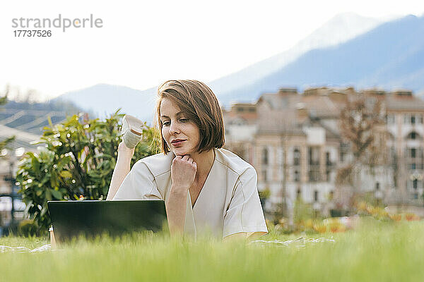 Frau mit Hand am Kinn und Laptop im Park liegend