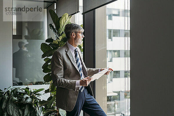 Geschäftsmann mit Tablet-PC und Dokumenten blickt durch Fenster im Büro