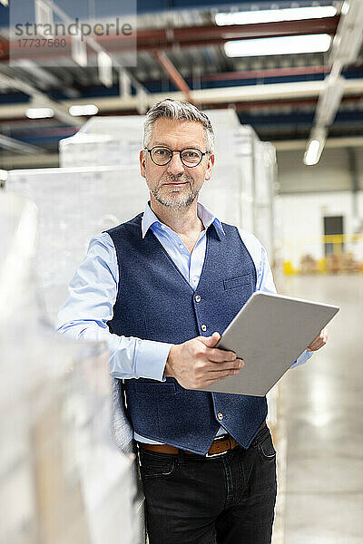 Manager mit Brille steht mit Tablet-PC neben Karton im Lager