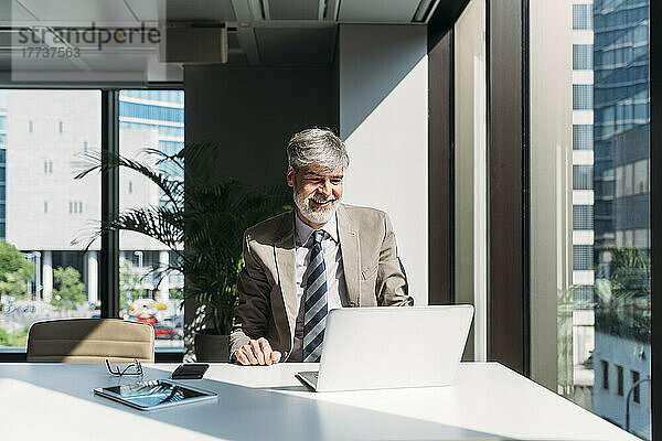Glücklicher Geschäftsmann mit Laptop  der am Fenster am Schreibtisch im Büro sitzt