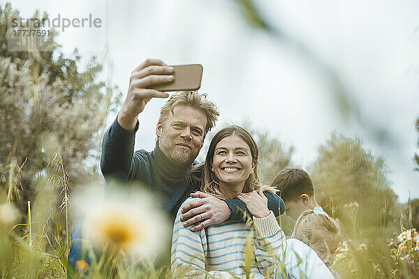 Smiling man with woman taking selfie through smart phone in meadow