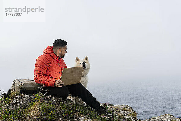 Man sitting with laptop looking at pet dog on rock