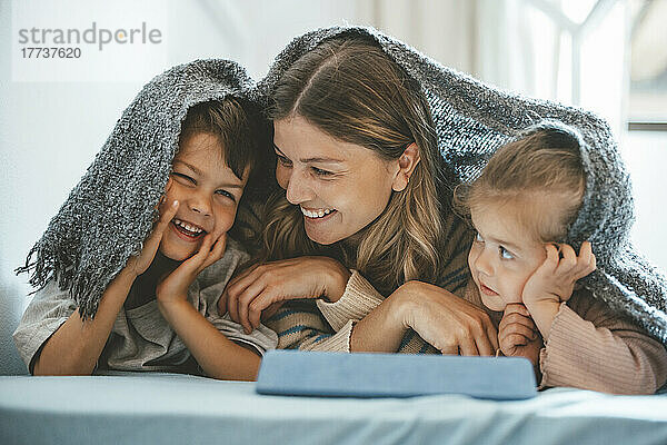 Lächelnde junge Frau und Mädchen schauen den Jungen an  der unter einer Decke auf dem Bett liegt