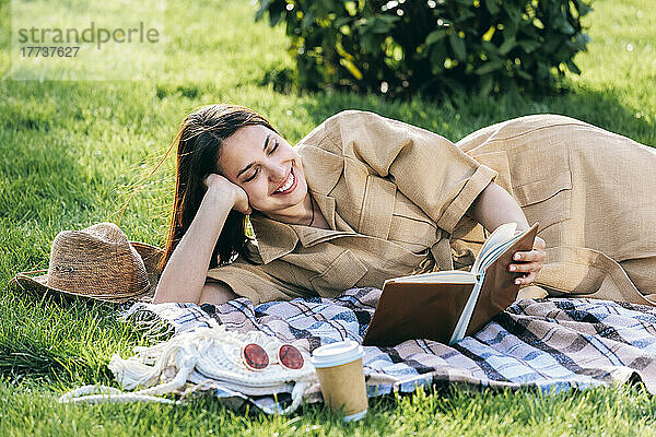 Glückliche junge Frau liest ein Buch und liegt auf einer Picknickdecke im Park