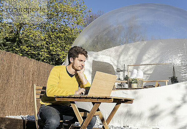 Young businessman using laptop sitting on chair in front of transparent dome hotel