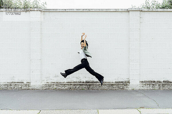 Cheerful young woman with arms raised jumping in front of wall