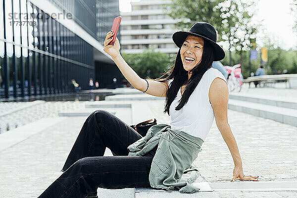 Fröhliche junge Frau mit Hut macht ein Selfie mit ihrem Smartphone und sitzt auf einer Treppe
