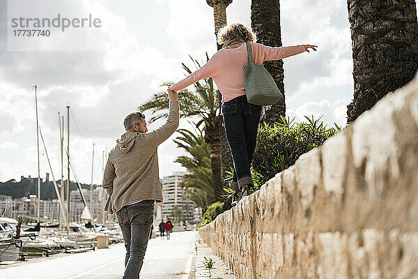 Reifer Mann hält die Hand einer Frau  die auf der Mauer am Hafen läuft