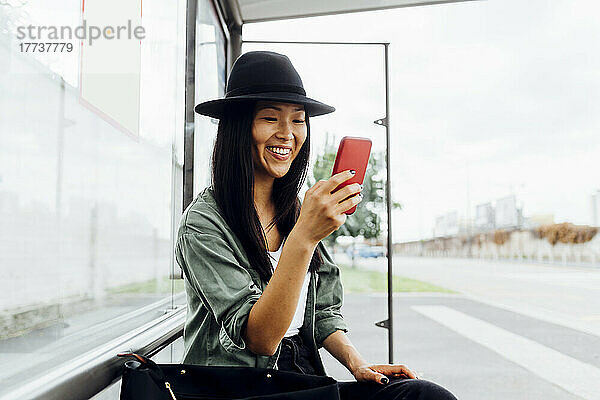 Lächelnde junge Frau surft per Smartphone im Internet und sitzt an der Bushaltestelle