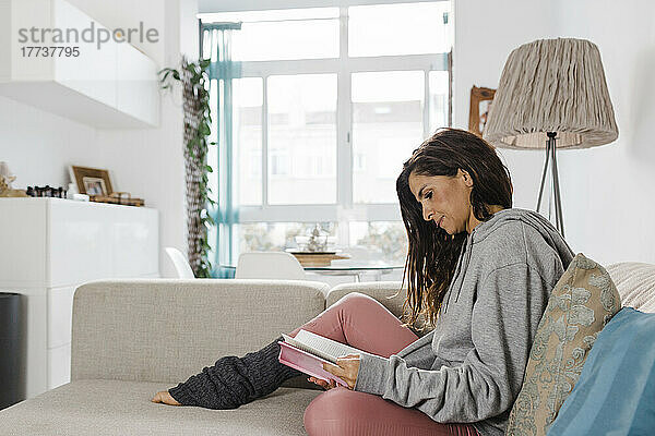 Reife Frau liest ein Buch und sitzt zu Hause auf dem Sofa im Wohnzimmer