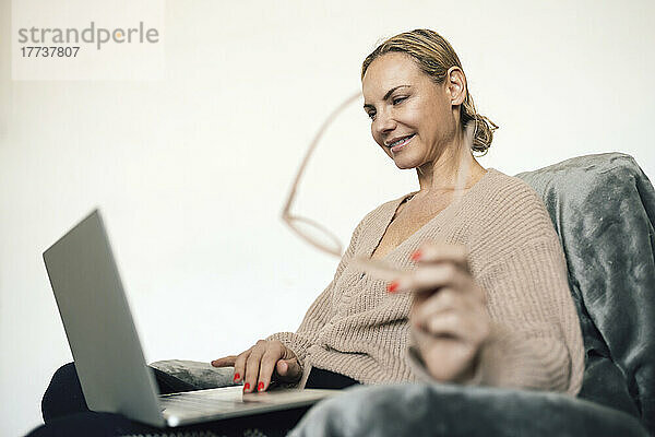 Lächelnde Geschäftsfrau mit Brille und Laptop auf dem Sessel zu Hause