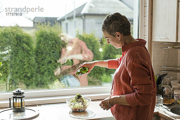 Frau am Fenster in der Küche gießt Olivenöl in eine Salatschüssel