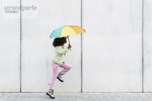 Mädchen genießt es  einen Regenschirm in der Hand zu halten und an einer weißen Wand vorbeizulaufen
