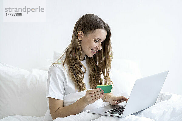 Glückliche Frau mit Kreditkarte beim Online-Einkauf per Laptop im heimischen Schlafzimmer