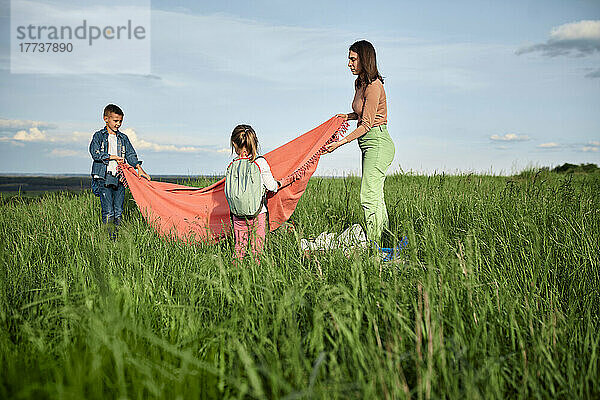 Mutter und Kinder legen Picknickdecke auf Gras im Feld aus