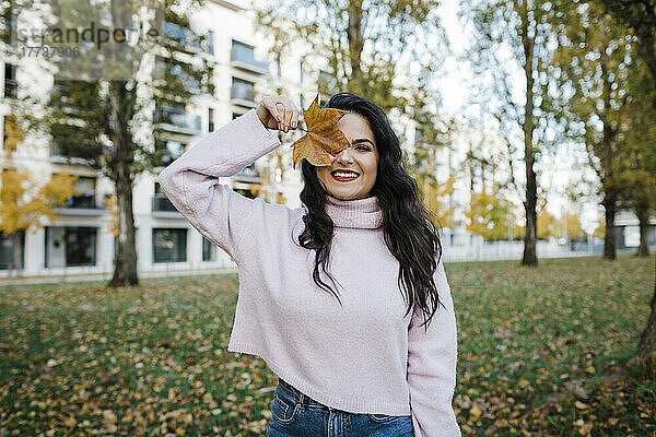 Glückliche junge Frau bedeckt Auge mit Herbstblatt im Park
