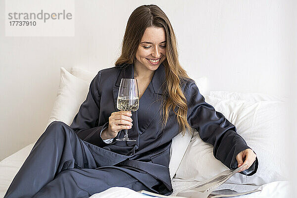Glückliche Frau mit Weinglas  die zu Hause im Bett eine Zeitschrift liest