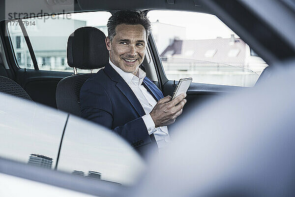 Lächelnder Geschäftsmann sitzt mit Smartphone im Auto