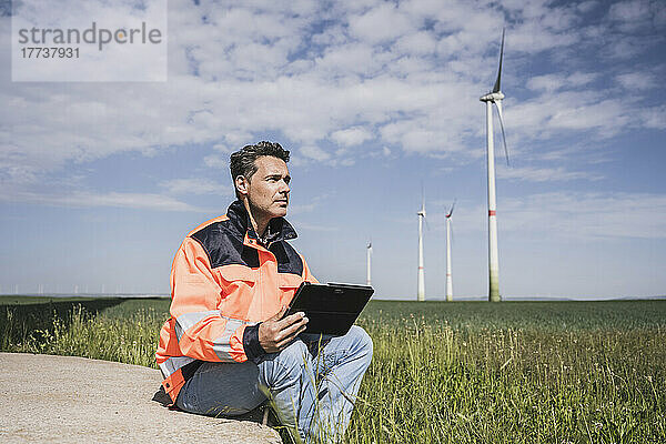 Ingenieur mit Tablet-PC sitzt neben Windkraftanlage auf dem Feld