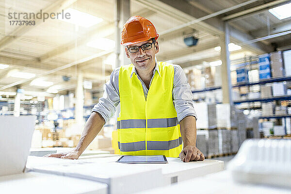 Lächelnder Arbeiter mit Brille lehnt an Karton im Lager