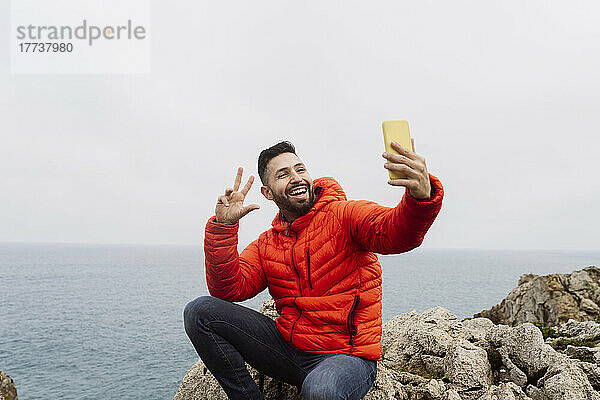 Glücklicher Mann gestikuliert Friedenszeichen und macht ein Selfie mit dem Smartphone  das auf einem Felsen sitzt