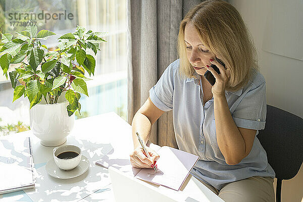 Blonde Frau telefoniert mit dem Handy und schreibt Tagebuch  sitzt am Schreibtisch im Heimbüro