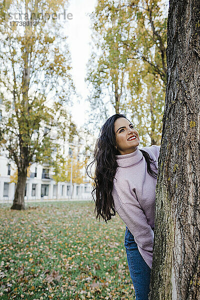 Glückliche junge schöne Frau  die den Herbst genießt und hinter einem Baumstamm im Park steht