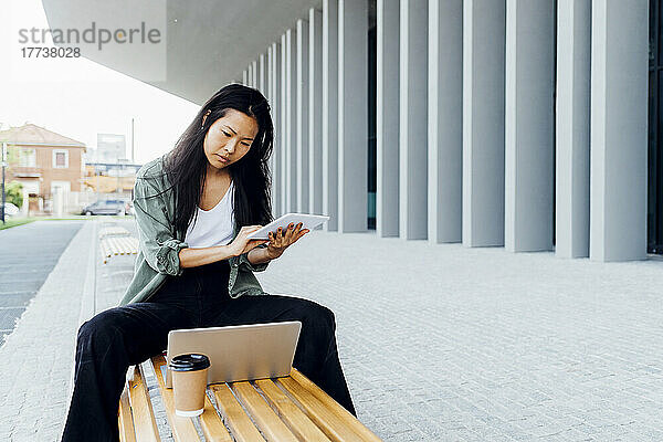 Junger Freiberufler mit Tablet-PC und Blick auf den Laptop auf der Bank