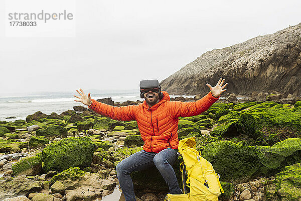 Glücklicher Mann mit Virtual-Reality-Simulator sitzt mit ausgestreckten Armen auf einem Felsen