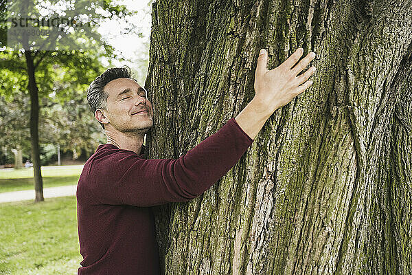 Lächelnder reifer Mann mit geschlossenen Augen umarmt Baum im Park