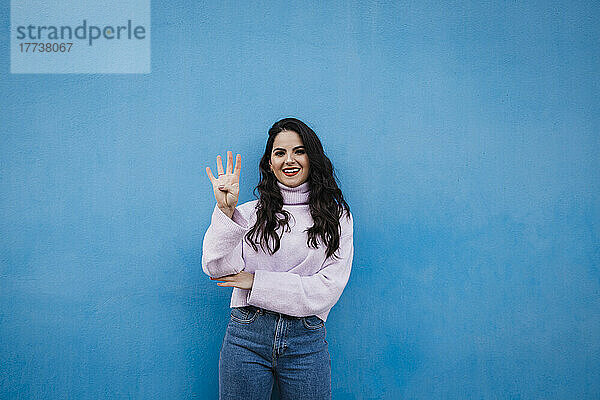 Lächelnde junge schöne Frau zeigt Nummer 4 vor blauer Wand