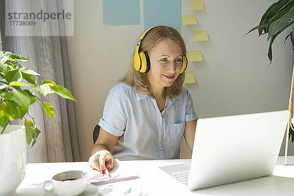 Lächelnder Freiberufler mit kabellosen Kopfhörern und Laptop am Schreibtisch im Heimbüro