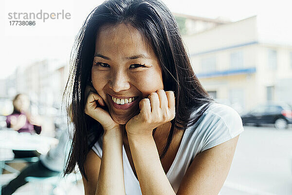 Glückliche junge Frau mit Kopf in den Händen sitzt im Straßencafé