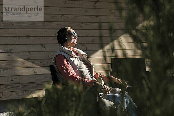 Frau mit Sonnenbrille und Kopfhörer sitzt im Sonnenschein auf der Veranda