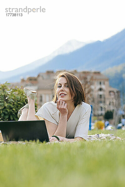Lächelnde Frau mit Laptop liegt im Gras im Park