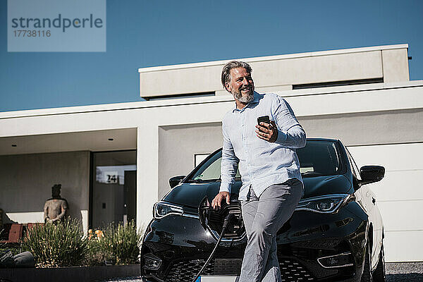 Lächelnder Mann hält an einem sonnigen Tag sein Mobiltelefon vor einem Elektroauto