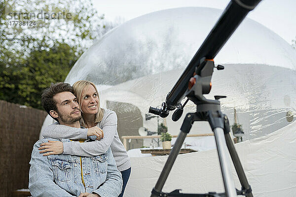 Frau umarmt Freund  der am Teleskop vor transparentem Kuppelhotel sitzt