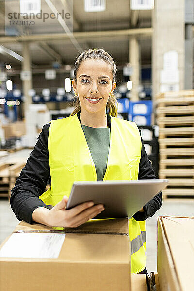 Lächelnder junger Arbeiter mit Tablet-PC steht neben Karton im Lager