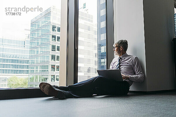 Geschäftsmann mit Laptop sitzt auf dem Boden am Fenster im Büro