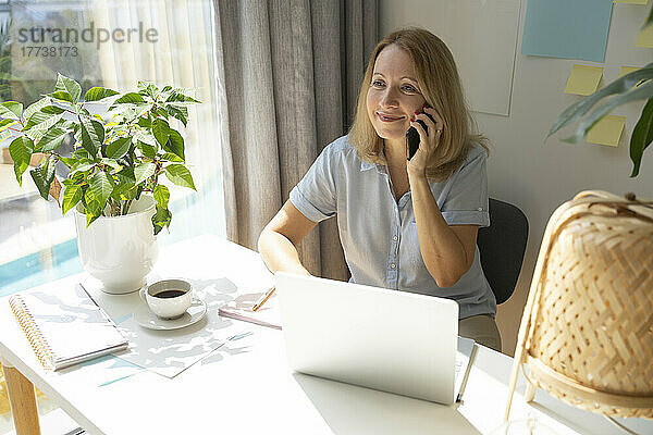Lächelnder blonder Freiberufler  der am Schreibtisch im Heimbüro sitzt und am Smartphone spricht