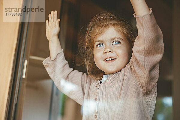 Happy cute girl at home looking out through glass window